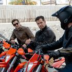 Drei Männer auf orangefarbenen KTM-Motorrädern, zwei in braunen Lederjacken, einer mit Helm. Sie schauen sich an.