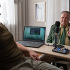 Zwei Personen sitzen an einem Tisch mit Laptop, Mikrofon und Podcast-Equipment. Eine trägt eine grüne Uniform.