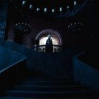 Keine lesbaren Texte. Dunkles, geheimnisvolles Interieur mit Treppe, Kronleuchtern und Person im Hintergrund.