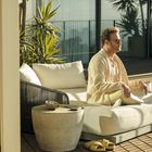 Ein Mann in beige Kleidung sitzt nachdenklich auf einem Liegesessel auf einer Terrasse, umgeben von Pflanzen und Sonnenlicht.