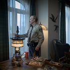 Ein Mann in einer Sheriff-Uniform steht in einem Raum, hält die Vorhänge. Dekor: Tisch mit Lampe, Wanddeko mit Vögeln.