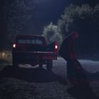 Ein Mann zieht eine Person auf einer dunklen, ländlichen Straße, nahe einem Pickup mit eingeschalteten Rücklichtern.