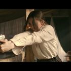 Eine Person in weißer Bluse zielt konzentriert mit einer Pistole, im Hintergrund Holzmöbel und große Fenster.