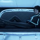 Eine Person in futuristischer, leuchtender Kleidung liegt entspannt auf einem transparenten Sofa in einem blau illuminierten Raum.