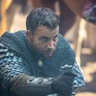 Ein Mann in mittelalterlicher Rüstung kniet mit ernstem Blick, hält ein Schwert. Hintergrund ist unscharf, Tiere und Menschen angedeutet.