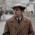 Ein Mann in einem braunen Mantel und Mütze steht auf einer belebten Straße. Im Hintergrund sind Menschen und Gebäude zu sehen.