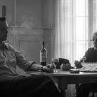 Zwei Männer an einem Tisch. Auf dem Tisch stehen Wein, ein Aschenbecher und Zigarettenpackungen. Rauch im Raum.