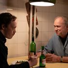 Zwei Männer sitzen in einer Küche an einem Tisch. Auf dem Tisch stehen Flaschen und sie scheinen im Gespräch vertieft.