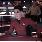 Eine Person in roter Uniform mit Anstecknadel auf einer futuristischen Brücke, umgeben von Displays und weiteren Crewmitgliedern.