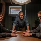 Drei Männer, mittleren Alters, am Tisch unter Lampe. Zwei sitzen, einer steht. Atmosphäre ernst, im Hintergrund Fenster.