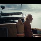 Text "TRADIESURF" auf einem Segel. Ein älterer Mann sitzt auf einem Boot, blickt aufs Meer, der Himmel ist bewölkt.