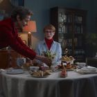 Drei Personen sitzen an einem Tisch mit Tee und Kuchen. Eine Person im roten Kleid reicht Blumen. Im Hintergrund Bücherregal.
