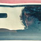 Keine Texte sichtbar. Eine Frau mit lockigem Haar und Sonnenbrille sitzt in einem Auto und schaut nach hinten.