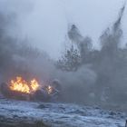 Ein umgekipptes Fahrzeug brennt, Flammen und Rauch steigen auf. Eine Person rennt in winterlicher Landschaft davon.