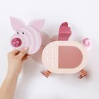 Eine Hand hält einen rosafarbenen, abstrakten Schweinekopf aus Papier mit Flügeln und Spiralschwänzchen.