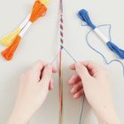 Zwei Hände knüpfen ein buntes Armband. Links orange-gelbes, rechts blaues Stickgarn.