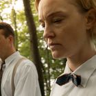 Zwei Personen in formeller Kleidung gehen durch einen grünen Wald, beide wirken nachdenklich und ernst.