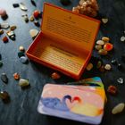 Offene orangefarbene Schachtel mit Text, umgeben von bunten Steinen und Karten mit Schwanenillustration.