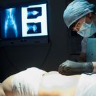 Chirurgin im OP, konzentriert arbeitend, neben ihr Röntgenaufnahmen, beleuchteter Raum, Patient auf OP-Tisch liegend.