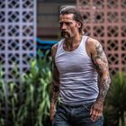 Ein Mann mit weißem Tanktop und Tattoos läuft draußen an einer gemusterten Mauer entlang.