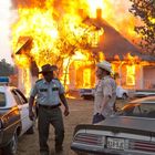 Nummernschild "Texas 518-BJE". Zwei Männer vor einem brennenden Haus, einer in Sheriff-Uniform, der andere mit Cowboyhut. Autos stehen daneben.
