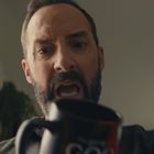 Die Tasse trägt den Text "GO". Ein Mann blickt intensiv darauf herunter, im Hintergrund sind ein Fenster und Vorhänge.