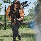 Ein Mann mit langen Haaren und bärtigem Gesicht trägt ein schweres Gewehr, rennt über eine Wiese, umgeben von Bäumen.