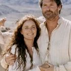 Ein Mann und eine Frau stehen in heller Kleidung lächelnd in einer sonnigen Landschaft, umarmt von anderen Menschen.