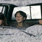 Vier Menschen sitzen in einem Auto, umgeben von grauer Asche. Sie blicken ernst durch die Windschutzscheibe.