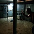 Zwei Menschen in einem dunklen, schmutzigen Raum. Eine sitzende Person blutverschmiert, eine andere steht mit Messer.