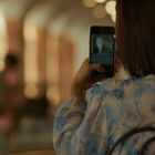 Eine Person fotografiert mit dem Handy, trägt eine blau-gemusterte Bluse, Umgebung ist unscharf und elegant beleuchtet.