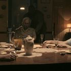 Personen sitzen an einem Tisch in einem spärlich beleuchteten Raum, eine trägt eine Sauerstoffmaske.