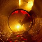 Keine erkennbaren Texte. Verschwommene Silhouette hinter beleuchtetem, nassem Tunnelgitter in orange-rotem Licht.