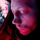 Ein Mann mit geschlossenen Augen, rotlich beleuchtet, sitzt in einer Umgebung mit verschwommenen Lichtern im Hintergrund.