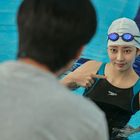 Eine Schwimmerin im Pool, trägt eine Badekappe und Schwimmbrille, zeigt auf sich selbst; blaues Wasser.