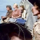 Vier Menschen auf einem Planwagen, einer trägt einen Cowboyhut und hat einen Schnurrbart. Sie sitzen zusammen, mit Blick in die Ferne.