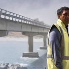 Ein Mann in gelber Warnweste steht nahe einer Brücke über einem Fluss, es ist windig.