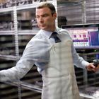 Ein Mann in weißer Schürze hält einen Scanner. Hinter ihm sind Regale und ein Bildschirm mit unleserlichem Text.