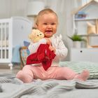 Ein fröhliches Baby in einem Raum hält ein Stofftier mit rotem Tuch. Im Hintergrund sind Gitterbett und Regal erkennbar.