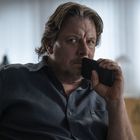 Ich kann keine Texte erkennen. Ein Mann sitzt nachdenklich und hält ein Smartphone vor seinen Mund. Im Hintergrund ist ein Büro.