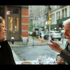 Ein älterer Mann und eine Frau sitzen an einem Tisch und unterhalten sich fröhlich in einem Café mit Blick auf die Straße.