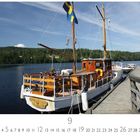 Mälarsee. Ein traditionelles Holzboot, mit schwedischer Flagge, liegt an einem Steg auf einem ruhigen See mit bewaldeten Ufern.
