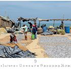 Fisch trocknet auf Jutematten unter einer einfachen Strandhütte, Menschen arbeiten, Meer im Hintergrund.