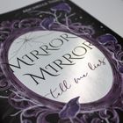 Text: "Mirror Mirror, tell me lies." Ovale Form mit violetten, floralen Mustern verziert.