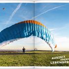 "Auszeit. Abenteuer. Lebensfreude." Ein bunter Paraglider über einer grünen Wiese vor blauem Himmel.