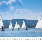 Zahl „9“ und Kalendertage unten. Segelboote auf ruhigem Wasser, Bäume und Berge im Hintergrund.