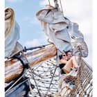 Text: "09". Zwei lächelnde Personen sitzen in einem Netz am Mast eines Segelschiffs, mit zusammengerollten Segeln darüber.