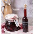 Text: Süße Rotwein-Zwetschken und Schwarzbeerlikör. Ein Glas voller Früchte, eine Flasche Likör auf einem Tuch.