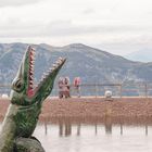 Grüner Dinosaurier mit offenem Maul vor Bergkulisse, Menschen spazieren auf einem Weg dahinter.