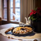 Ein Teller mit Keksen auf einem Tisch mit weißem Häkeldeckchen, daneben ein Kännchen-Set und eine Weihnachtssternpflanze.